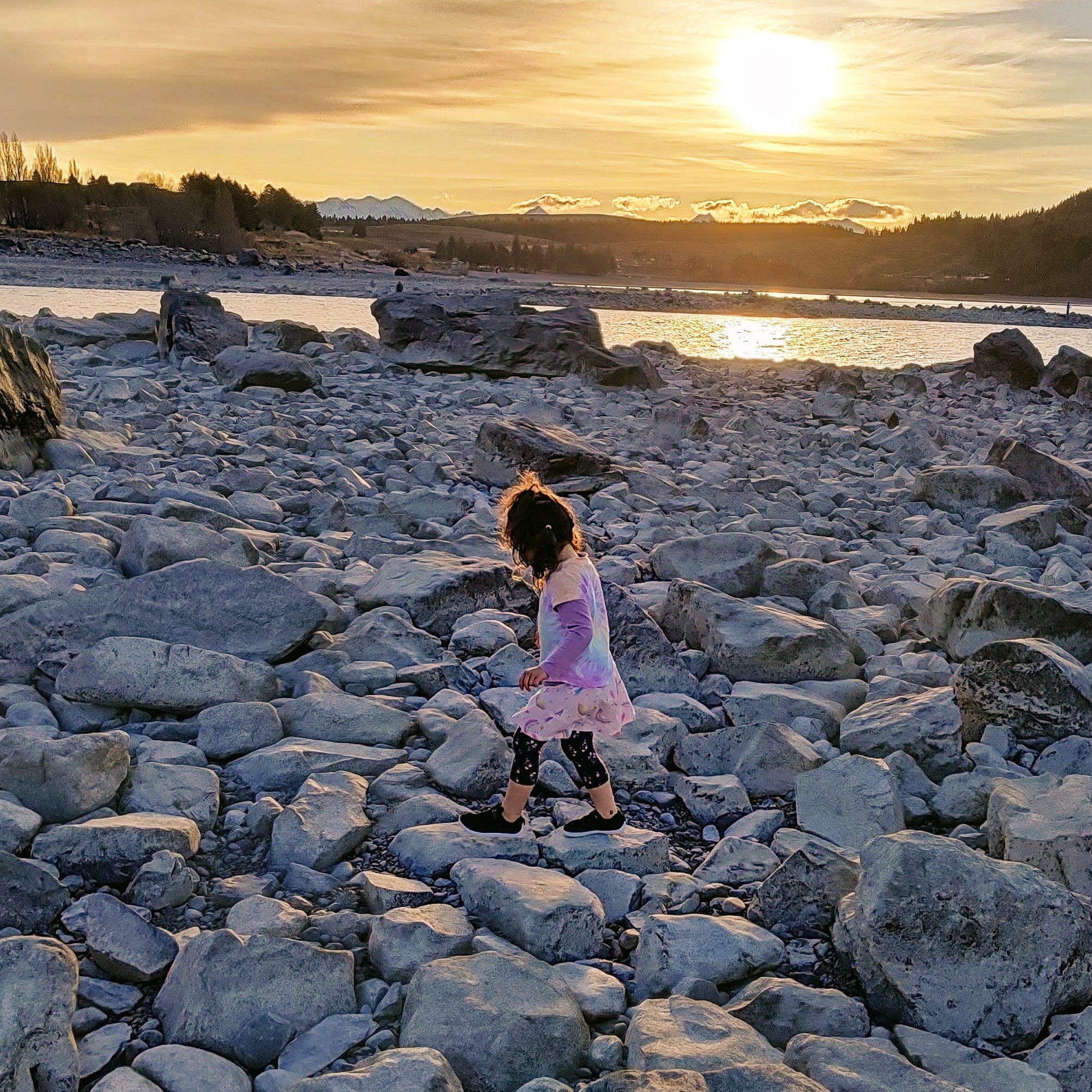 Person standing on a rocky shore at sunset with mountains in the background