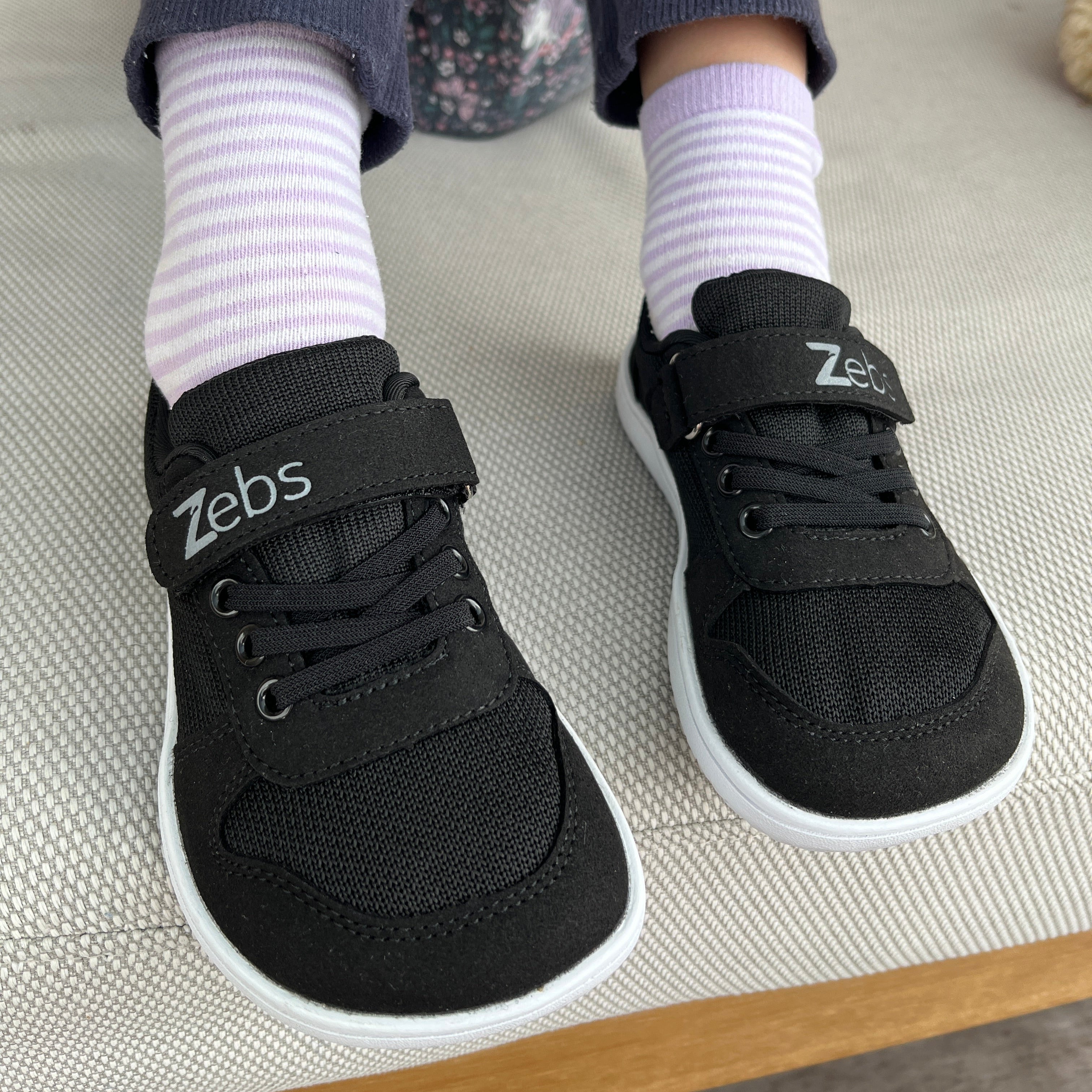 Black sneakers with 'zebs' branding worn by a person on a light-colored couch.